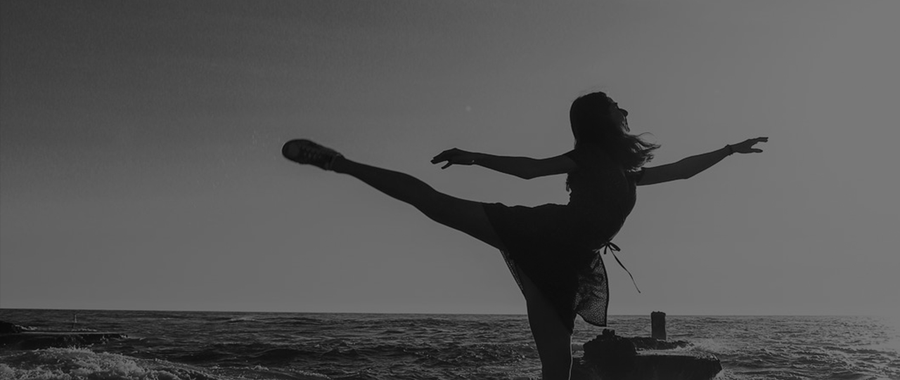 Woman in flowy linen wrap dress and sneakers standing by ocean in first arabesque ballet position